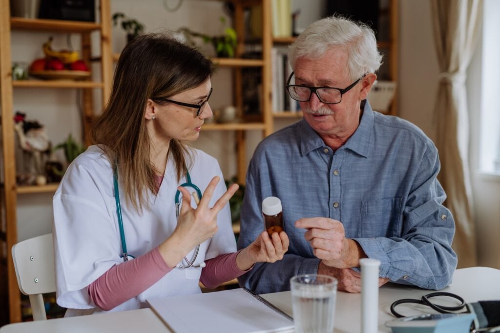 Healthcare worker or caregiver visiting senior man indoors at home, explaining medicine dosage.