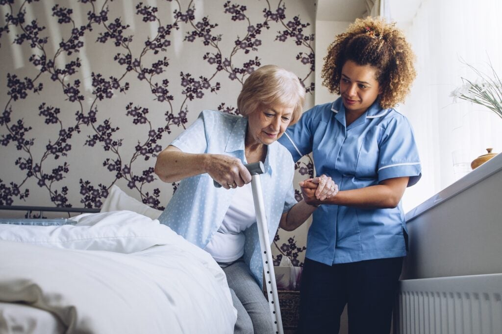 Home Caregiver helping a senior woman get up from her bed with a walking stick.