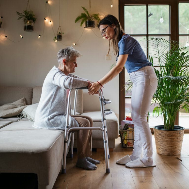 A homecare assistant helping a woman stand up from a sofa using a walker
