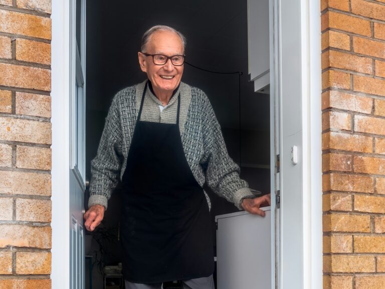 An elderly man dressed in a kitchen apron smiling in the doorway of a house