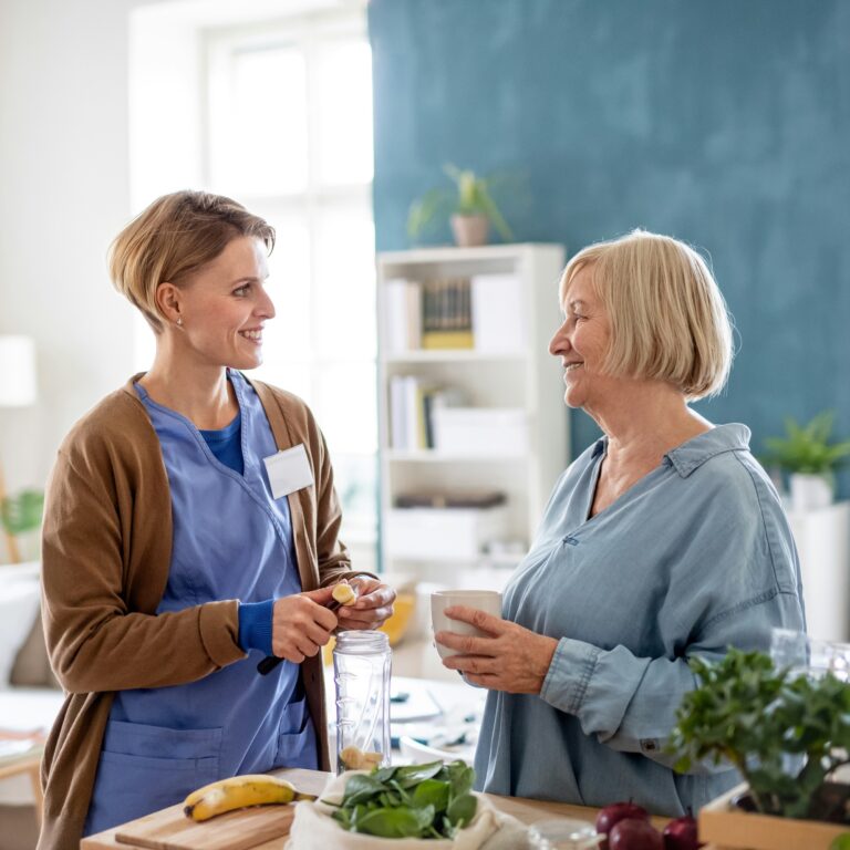 A woman and a nurse talking as the nurse slices bananas into a blender