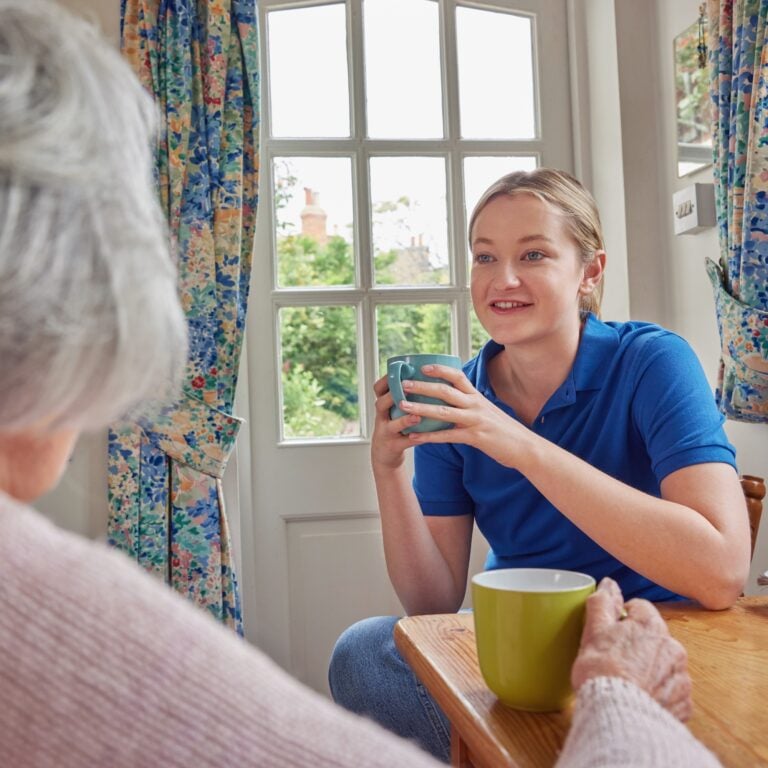 Female Home Help Having Cup Of Tea With Lonely Senior Woman In Kitchen