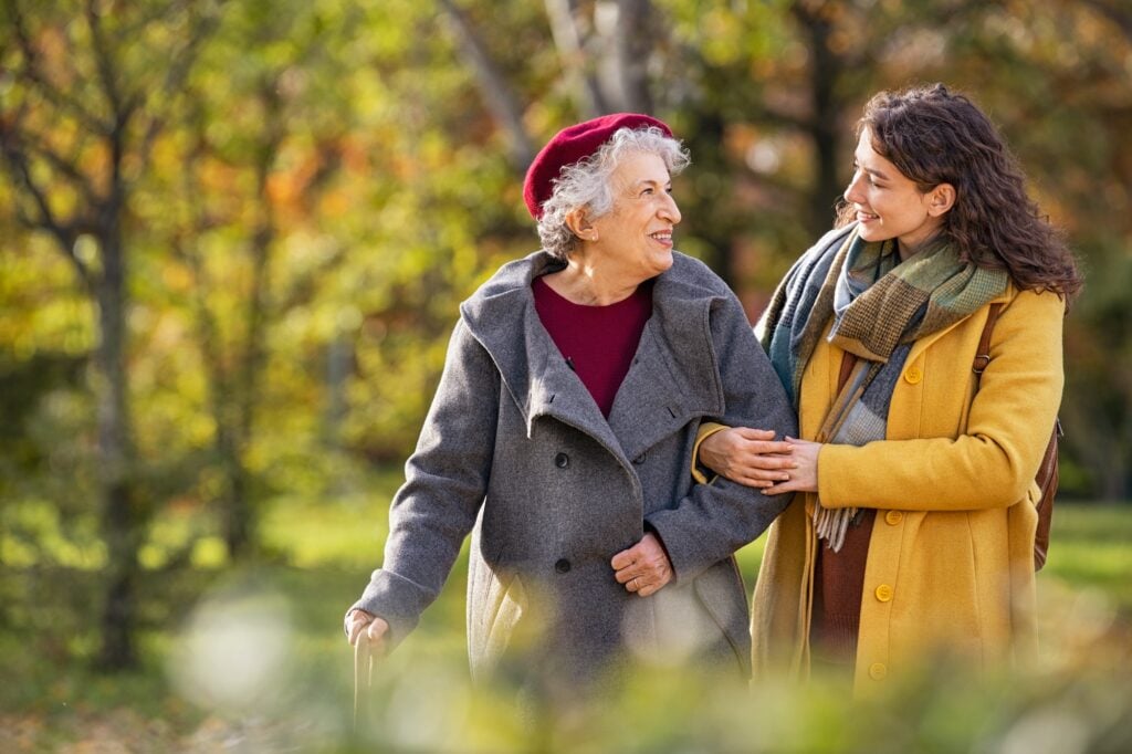 Young woman in park wearing winter clothing walking with old grandmother. Smiling lovely caregiver and senior lady walking in park during autumn and looking at each other with copy space.