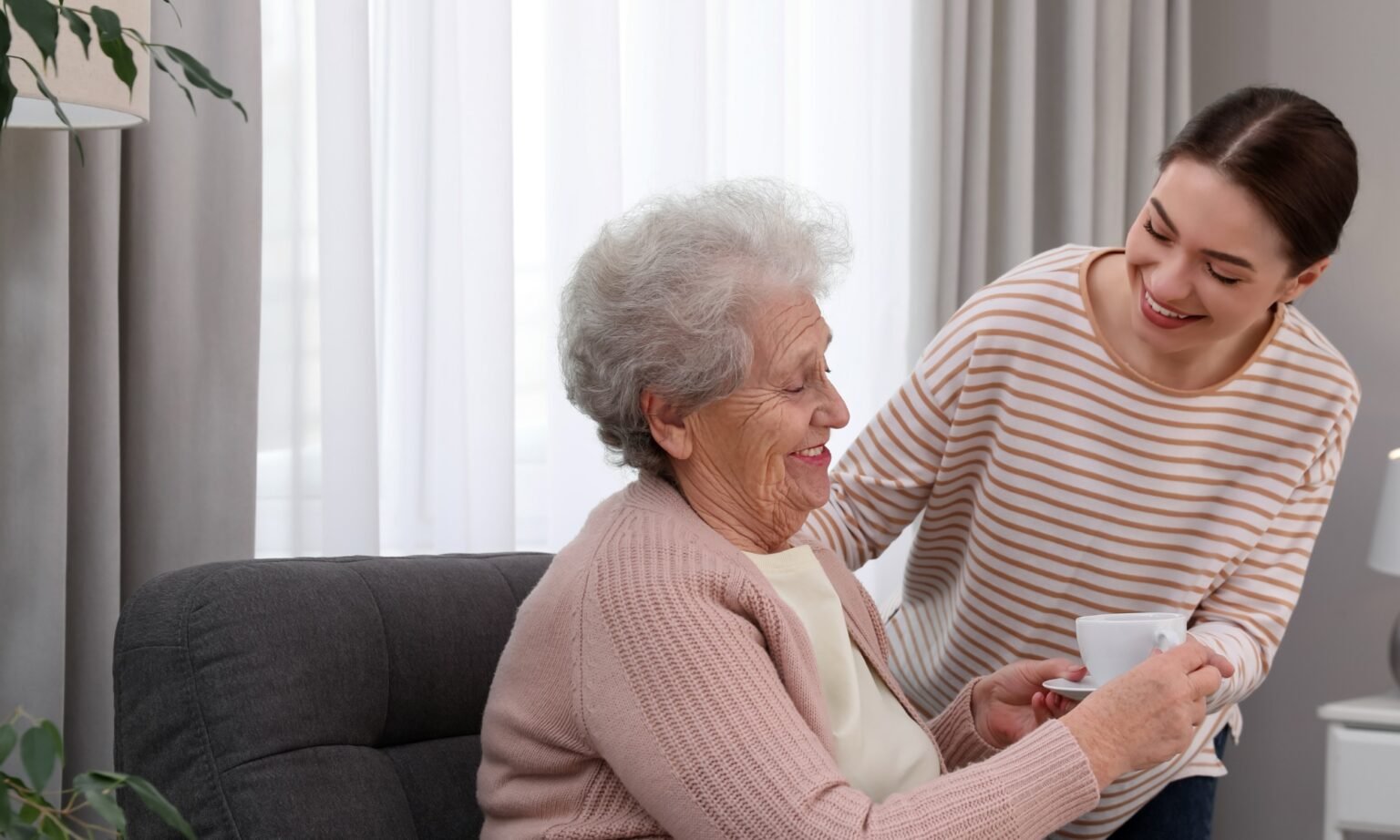 Young caregiver giving tea to senior woman in room. Home care service