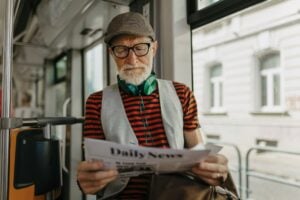 Elderly man traveling through the city by bus, reading newspaper. Senior city commuter taking tram to grocery, using public transportation.