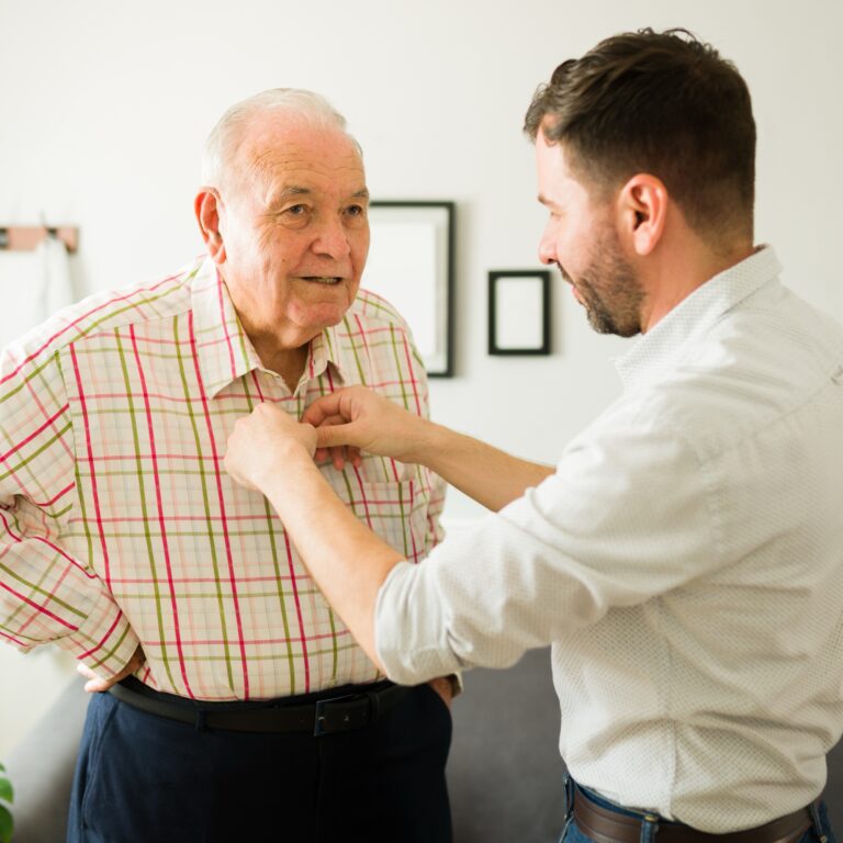 Caring young son helping his old happy elderly father to change clothes before going out of the house together.